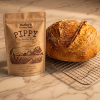 A kraft paper pouch labeled 'Mother's Country Store Pippy The Starter Fuel' stands beside a round artisanal bread loaf on a metal cooling rack over a marble surface.