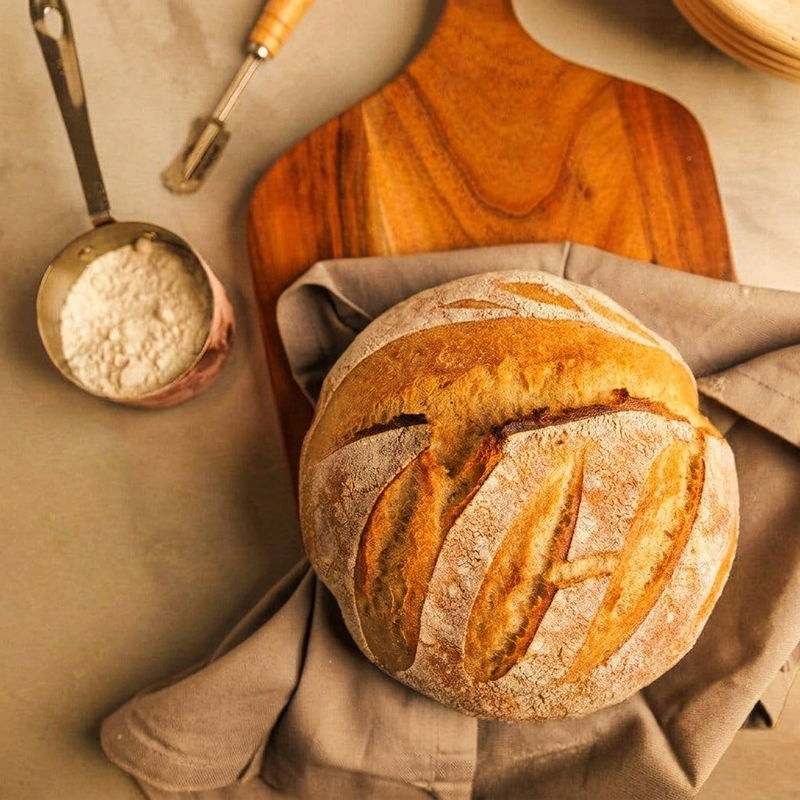 A round artisanal loaf of bread with a golden-brown crust and flour-dusted scoring, resting on a beige cloth on top of a wooden cutting board. A copper measuring cup filled with flour sits to the left on a neutral beige surface.