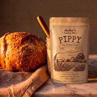A stand-up pouch labeled 'Pippy The Starter Fuel' by Mother's Country Store stands on a rustic wooden surface beside a loaf of bread and a small jar with a wooden spoon, in warm studio lighting.